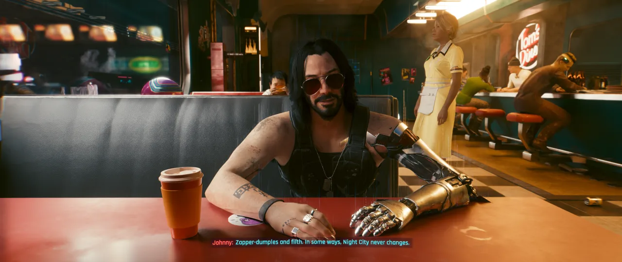 A seated character with long dark hair, sunglasses, and a visible cybernetic arm leans across a table in a warm, neon-lit interior, with a drink cup in the foreground and other figures blurred in the background. The scene fits the *Cyberpunk 2077* gallery’s conversational, story-focused tone, capturing a relaxed but tense moment inside a sleek futuristic space.