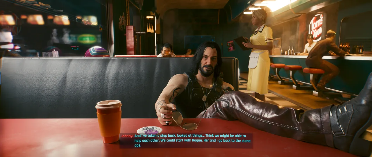 A relaxed interior scene from *Cyberpunk 2077*, showing a man reclining with his feet up in a warmly lit bar or lounge while another figure stands in the background near the counter. The shot emphasizes the game's stylish, lived-in spaces and dialogue-driven atmosphere.