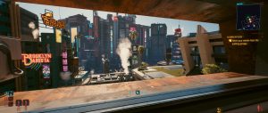 A wide *Cyberpunk 2077* city view fills the frame, seen through a large interior window that opens onto dense futuristic high-rises, glowing signs, and drifting smoke in warm late-day light. The scene blends a sleek apartment-like setting with the game’s HUD, emphasizing the contrast between the calm room and the busy skyline beyond.