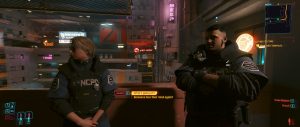 A dialogue scene from *Cyberpunk 2077* shows two characters facing each other inside a compact, neon-lit interior, with one in an NCPD-style uniform and the game’s conversation interface visible across the screen. The warm lighting, cluttered tech-filled room, and muted city glow in the background give the moment a tense, story-focused feel.