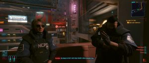 A dialogue scene from *Cyberpunk 2077* shows two characters seated in a dim, neon-lit interior, with one wearing an NCPD shirt while the other sits in darker clothing across the room. The frame emphasizes the game’s moody, futuristic atmosphere through its warm signage, cluttered urban backdrop, and visible conversation UI.