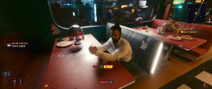 A *Cyberpunk 2077* dialogue scene shows a man seated alone at a wide red table in a sleek, neon-lit interior, with food, bottles, and scattered items around him while the game’s conversation UI remains visible on screen.