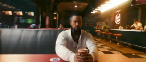 A bearded man in a white jacket sits alone at a diner-style counter, holding a cup as warm neon lighting and a checkerboard floor give the *Cyberpunk 2077* scene a moody, late-night feel.