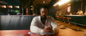 A seated man in a white jacket rests his hands on a table in a softly lit diner-like interior, with warm reflections, neon accents, and other patrons visible along the counter behind him. The framing gives the scene an intimate, dialogue-focused *Cyberpunk 2077* feel.