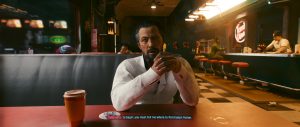 A man in a white shirt sits at a table in a softly lit futuristic diner or bar, holding a cup as he looks ahead, with another drink set in the foreground and the warm, neon-tinged interior stretching out behind him.