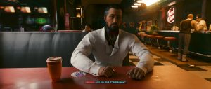 A bearded man in a white shirt sits across a wooden table in a warmly lit bar or diner, with a takeaway coffee cup and small items in the foreground while other patrons linger in the background. Framed like a dialogue scene from *Cyberpunk 2077*, the image focuses on a calm, story-driven conversation rather than action.