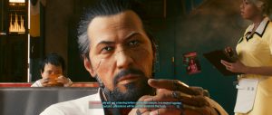 A close-up dialogue scene from *Cyberpunk 2077* shows Takemura seated indoors, holding a cigarette while speaking, with other figures and warm ambient lighting in the background. The framing emphasizes the conversation and the game’s cinematic, story-focused atmosphere.