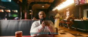 A seated man in a bright, modern diner-style interior appears mid-conversation, framed by warm overhead lighting, glossy counters, and a few other patrons in the background. As part of this *Cyberpunk 2077* gallery, the scene emphasizes a quieter, dialogue-focused moment rather than action.