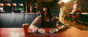 A seated character with long dark hair, sunglasses, and a visible cybernetic arm leans across a table in a warm, neon-lit interior, with a drink cup in the foreground and other figures blurred in the background. The scene fits the *Cyberpunk 2077* gallery’s conversational, story-focused tone, capturing a relaxed but tense moment inside a sleek futuristic space.