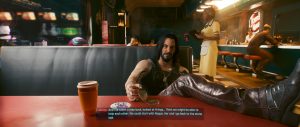 A relaxed interior scene from *Cyberpunk 2077*, showing a man reclining with his feet up in a warmly lit bar or lounge while another figure stands in the background near the counter. The shot emphasizes the game's stylish, lived-in spaces and dialogue-driven atmosphere.