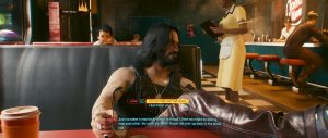 A *Cyberpunk 2077* dialogue scene set in a warmly lit diner or bar, showing a long-haired character seated in a booth with a drink in the foreground while the game’s conversation interface and subtitles frame the moment.