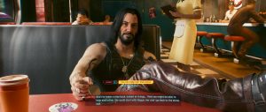 A relaxed conversation scene from *Cyberpunk 2077*: a long-haired character lounges across a diner booth with a drink on the table, while the warm, busy interior and on-screen dialogue UI give the moment a casual, story-driven feel.