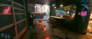 A dim, futuristic interior from *Cyberpunk 2077* shows a cluttered room with glowing floor lights, mesh partitions, and a long desk set beneath bright screens and window light, capturing the game’s gritty high-tech atmosphere during a quieter story moment.